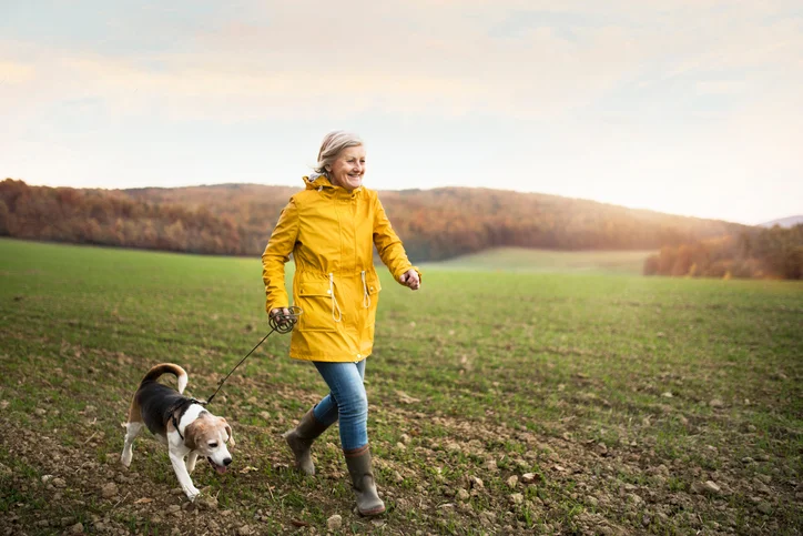 Woman in yellow coat walking a dog in a field during autumn.