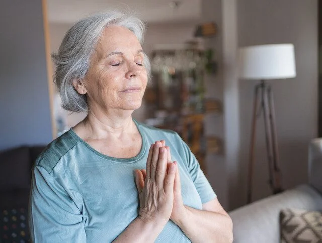 Elderly woman meditating with eyes closed, hands in prayer position, in a cozy living room.