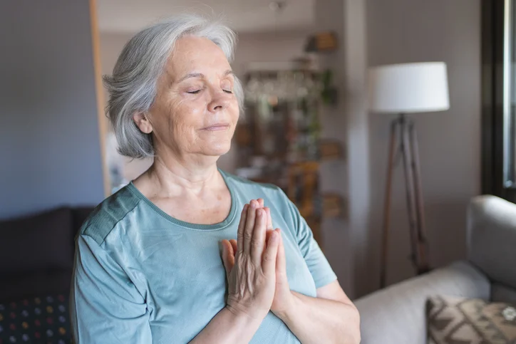 Elderly woman meditating with eyes closed, hands in prayer position, in a cozy living room.