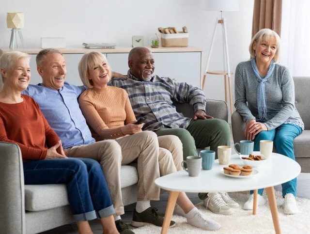 Seniors smiling and sitting on a couch with coffee and snacks on the table.