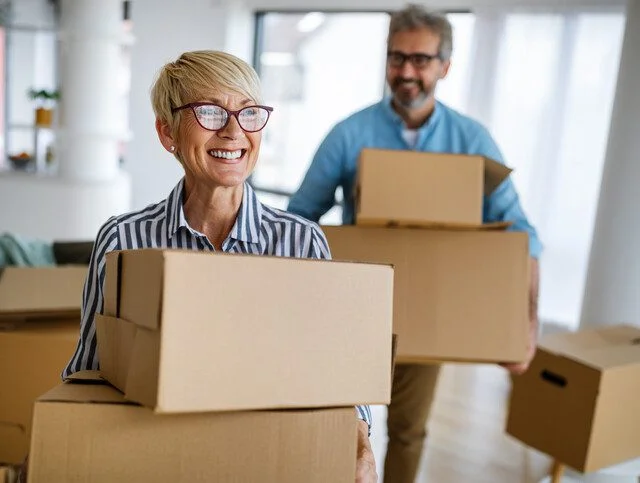 Smiling woman and bearded man move boxes in a well-lit room.