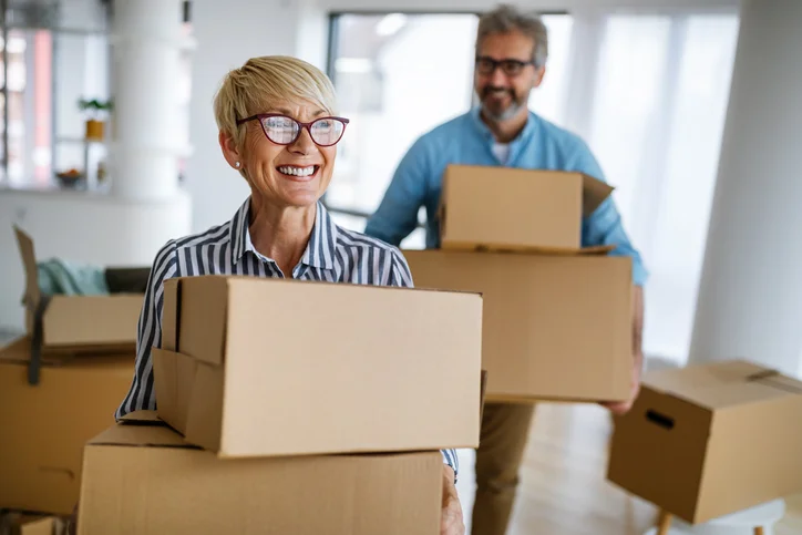 Smiling woman and bearded man move boxes in a well-lit room.