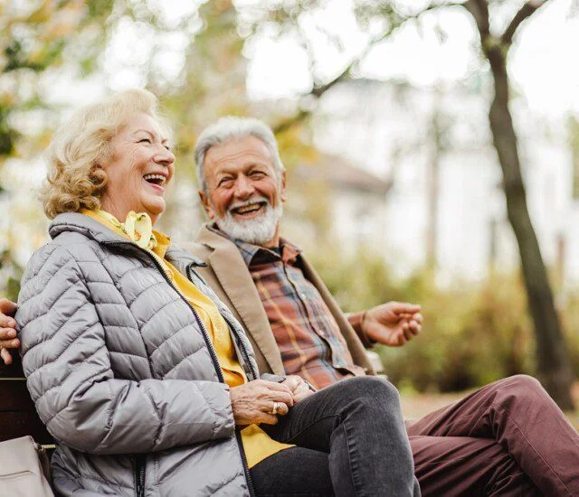 Elderly couple laughing together on a park bench, surrounded by trees.