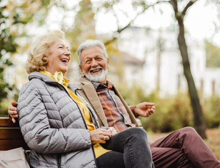 Elderly couple laughing together on a park bench, surrounded by trees.