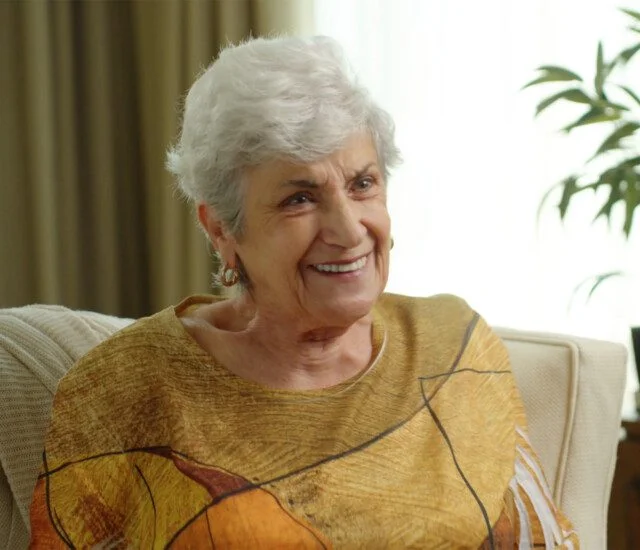 Smiling elderly woman seated on a beige chair in a warmly lit room.