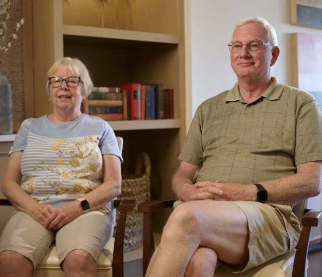 Older couple sitting and smiling in a cozy room with bookshelves.