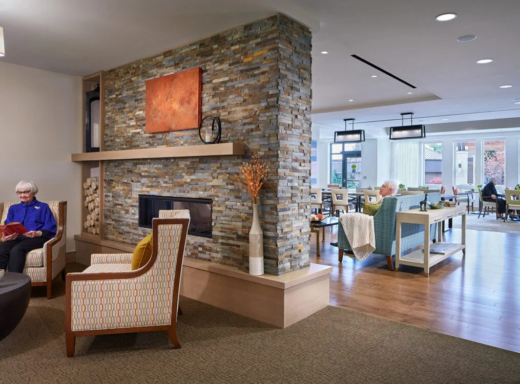 Spacious lounge with seniors seated, featuring a stone fireplace, modern furniture, and large window