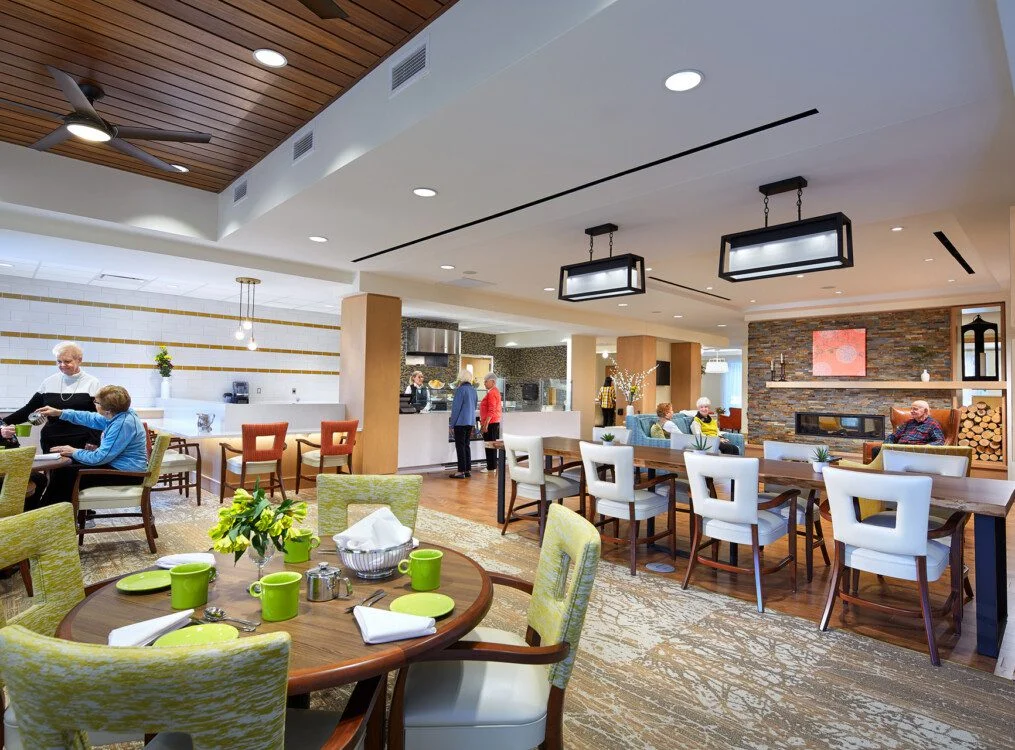 Modern community lounge with seating, dining tables, and a fireplace. People are socializing.