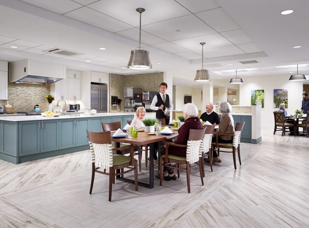 Dining area with seniors being served, modern kitchen in background.