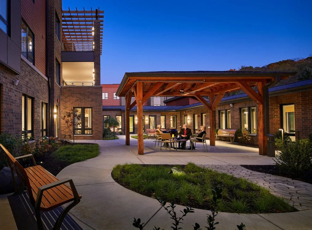 Courtyard with benches and people at a table under a wooden pergola at dusk.