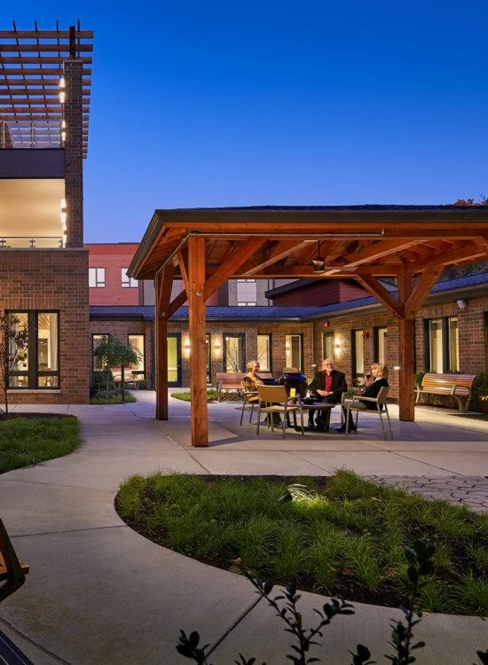 Courtyard with benches and people at a table under a wooden pergola at dusk.