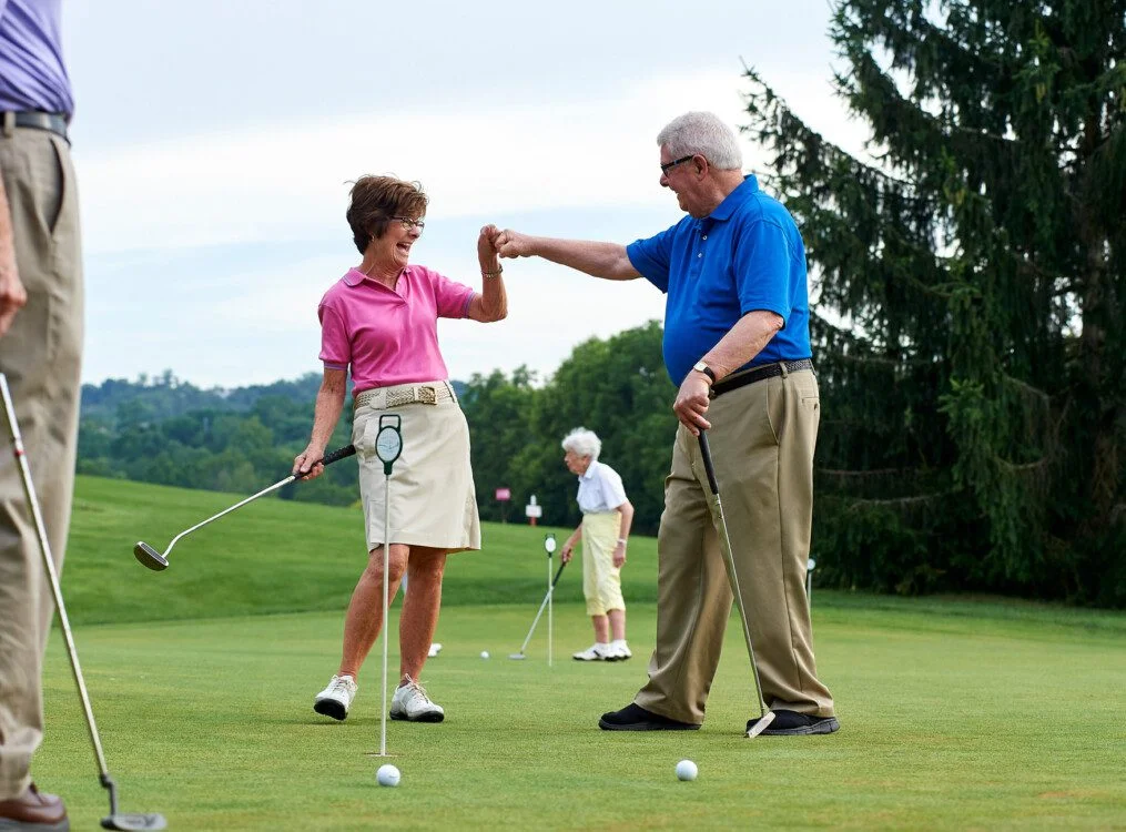 Two people share a fist bump on a golf course, holding clubs, with others in the background.