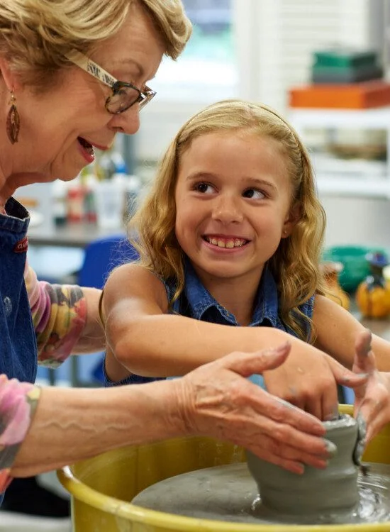 Elderly woman teaching girl pottery on a wheel, both smiling in a classroom.