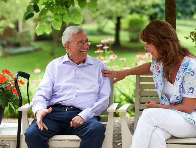 Elderly man and woman smiling on a bench in a garden setting.