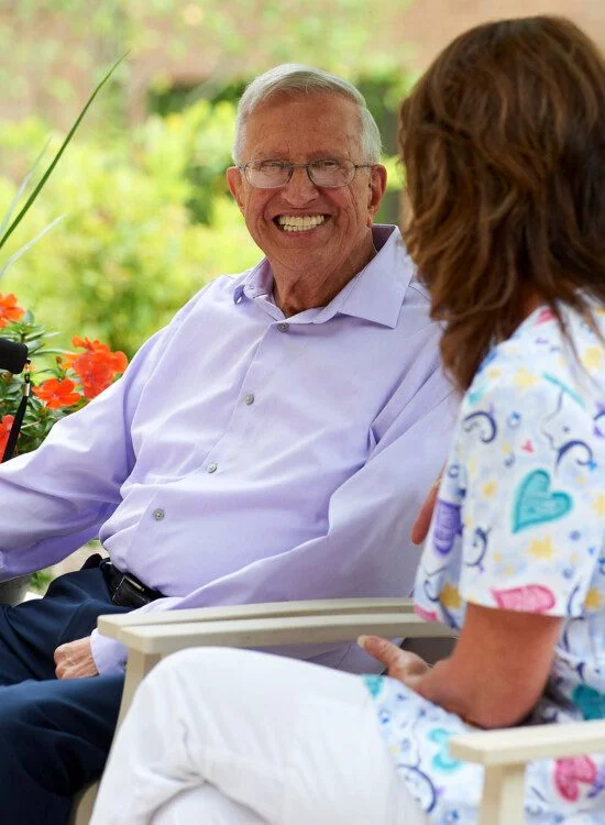 Elderly man and woman chatting outdoors, both smiling warmly.
