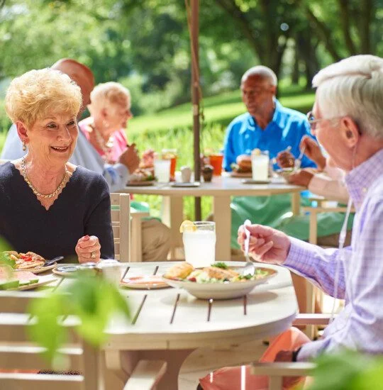 Seniors enjoying a meal together outdoors at a garden patio on a sunny day.