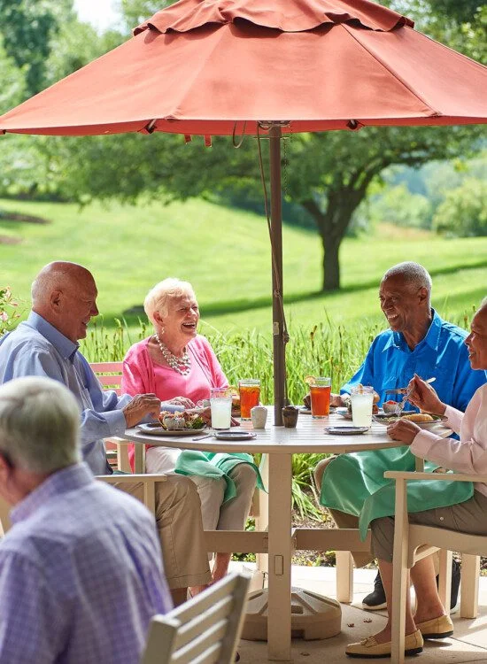 Seniors enjoy a meal outdoors under an umbrella on a sunny day.