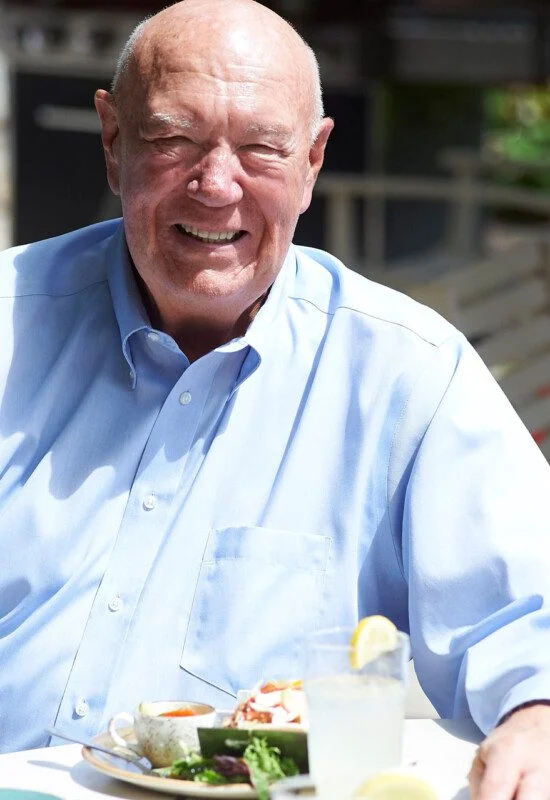 Smiling man in a blue shirt dining outdoors at a table with food and drinks.