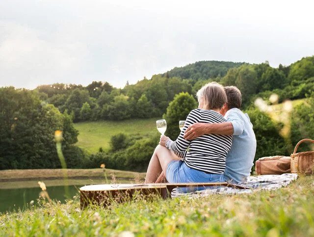 Couple enjoys wine at picnic by lake, surrounded by trees and hills.