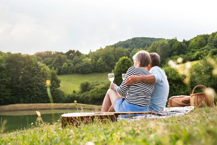 Couple enjoys wine at picnic by lake, surrounded by trees and hills.