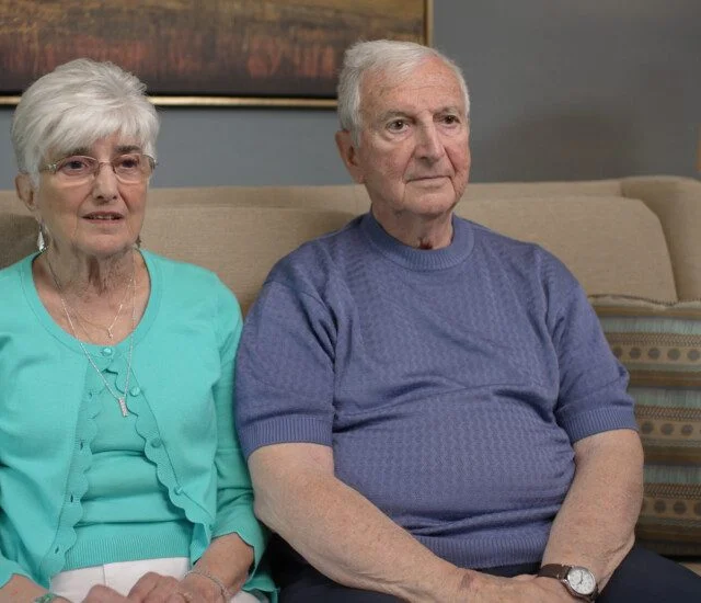 Elderly couple sitting on a sofa, wearing blue and turquoise tops, with a lamp in the background.