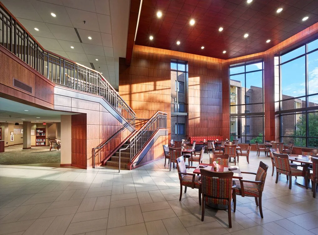 Spacious lobby with wooden walls, large windows, tables, and staircases.