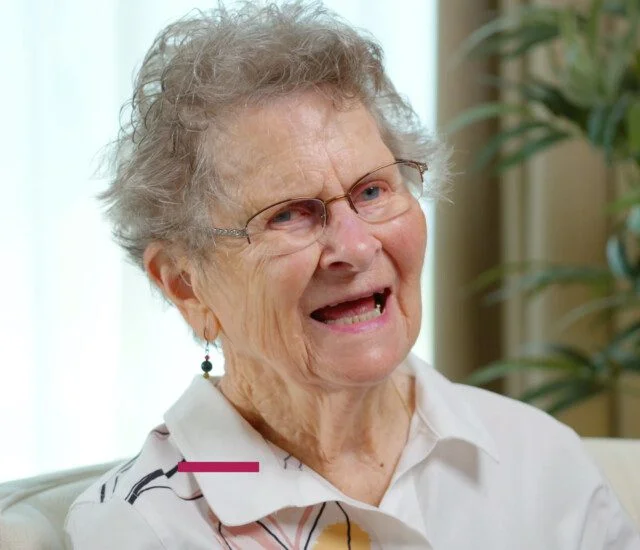 Elderly woman smiling indoors, wearing glasses and earrings.