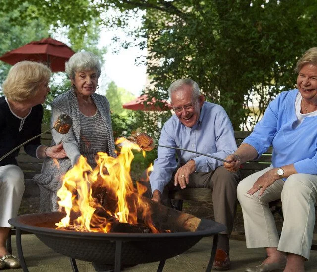Four seniors roasting marshmallows over a fire pit, smiling and enjoying the outdoors.