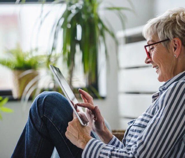Older woman using a tablet, sitting in a bright room with plants.