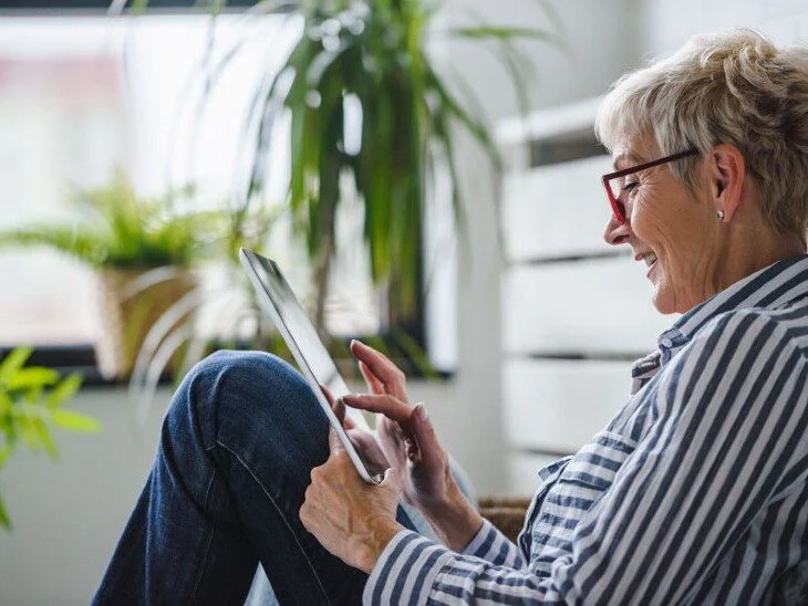 Older woman using a tablet, sitting in a bright room with plants.