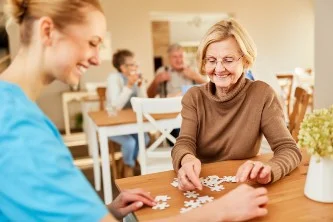 Elderly woman and caregiver smiling while assembling a puzzle at a table.