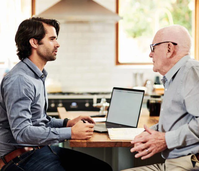 Two people having a discussion at a table with a laptop and papers.