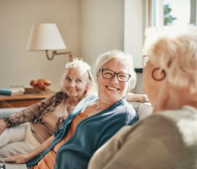 Three elderly women smiling and talking on a couch in a bright room.