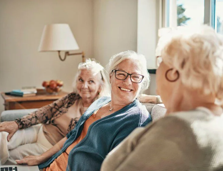Three elderly women smiling and talking on a couch in a bright room.