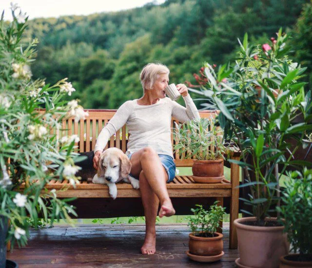 Woman drinking coffee on a patio bench with a dog, surrounded by potted plants.
