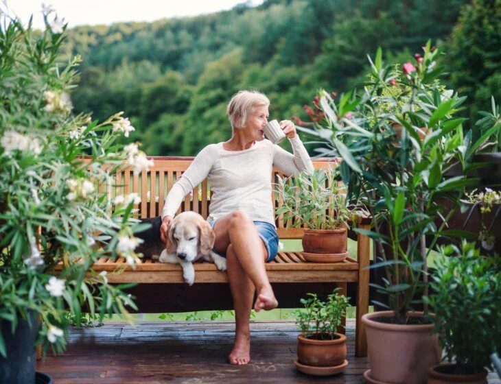 Woman drinking coffee on a patio bench with a dog, surrounded by potted plants.