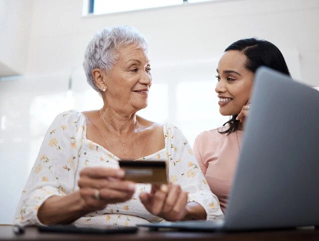 Two women using a laptop and credit card, smiling at each other.