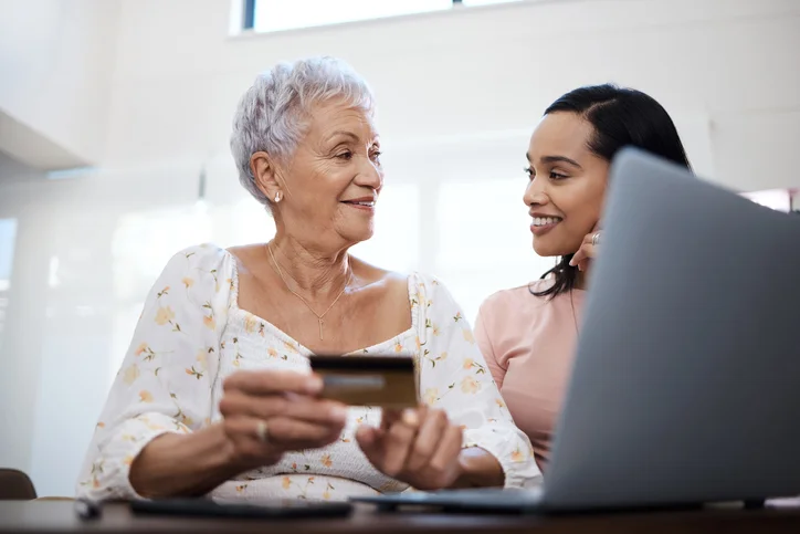 Two women using a laptop and credit card, smiling at each other.
