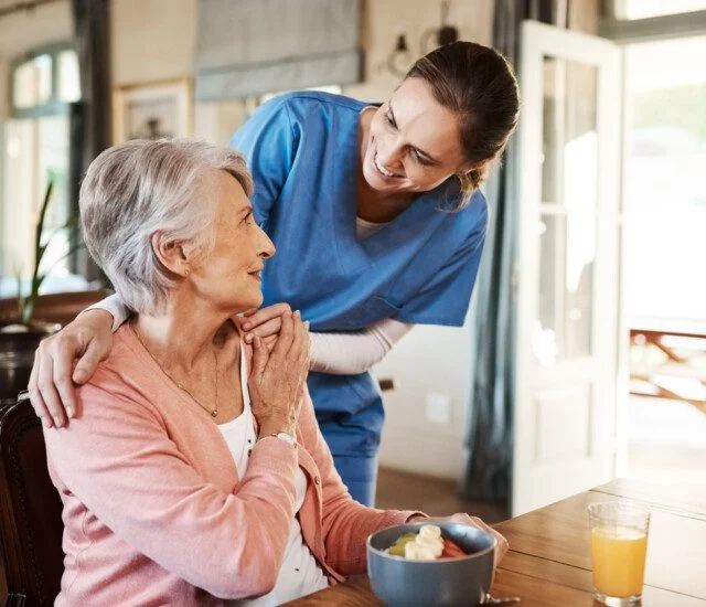 Caregiver in blue uniform assisting a smiling senior woman at a dining table.