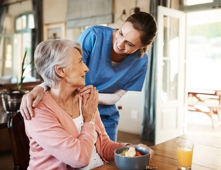 Caregiver in blue uniform assisting a smiling senior woman at a dining table.