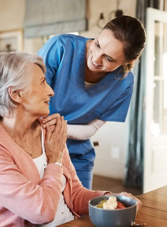 Nurse smiling at elderly woman sitting at a table with a meal.