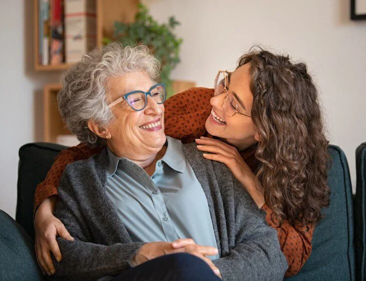 Elderly woman and younger woman smiling together on a couch.