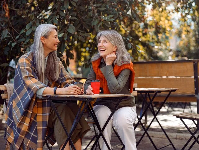 Two women sit outdoors, enjoying coffee and chatting at a wooden table.