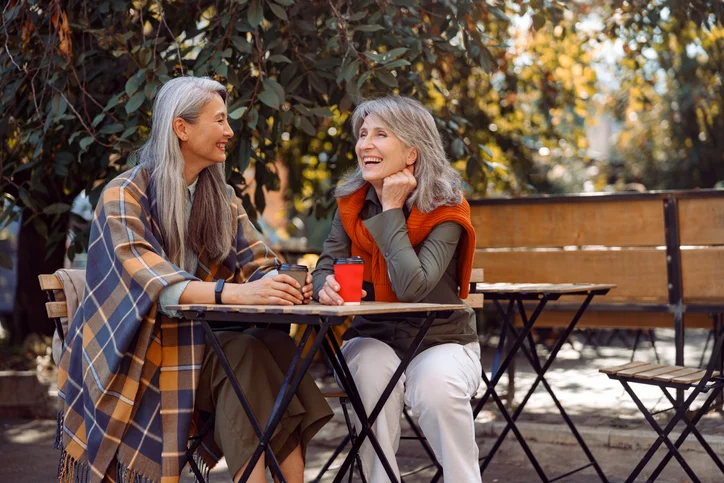 Two women sit outdoors, enjoying coffee and chatting at a wooden table.