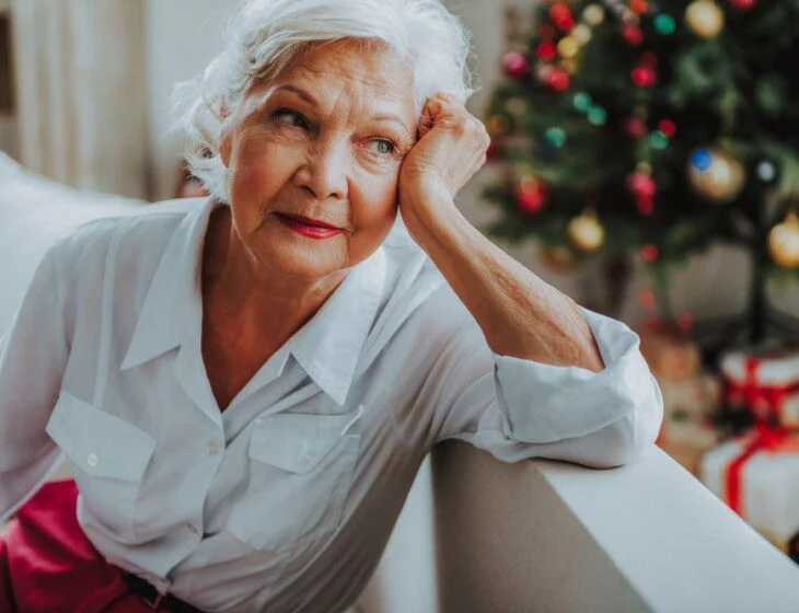 Elderly woman in white blouse sitting pensively near a decorated Christmas tree.