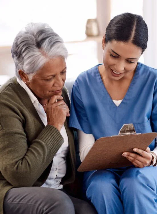 Nurse in blue scrubs discussing a document with an older woman on a couch.