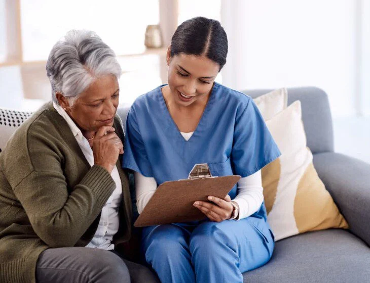 Nurse showing an elderly woman a clipboard while sitting on a sofa.