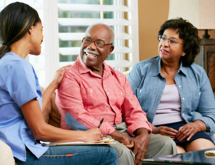 Caregiver talking with an elderly couple, smiling indoors.