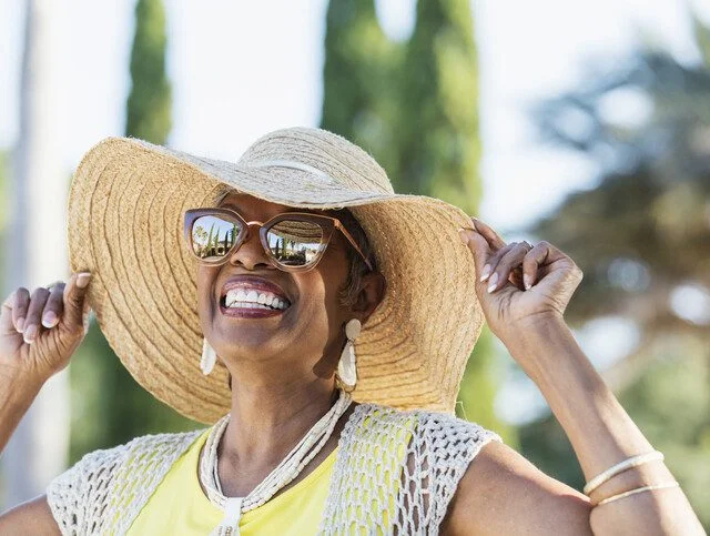 Smiling woman in sun hat and sunglasses outdoors on a sunny day.