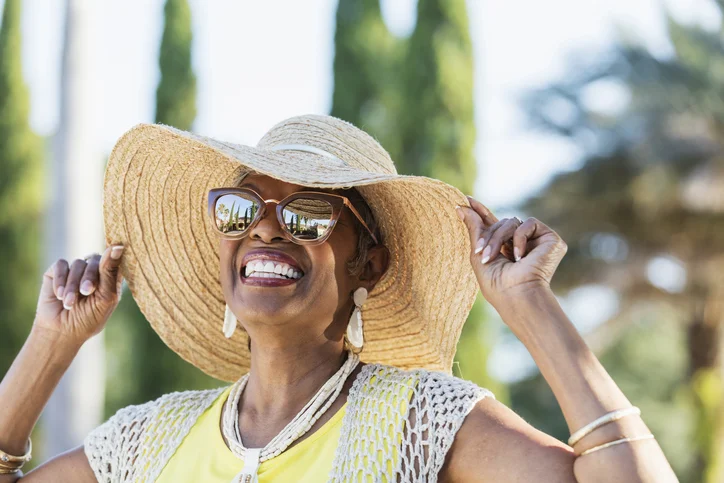 Smiling woman in sun hat and sunglasses outdoors on a sunny day.